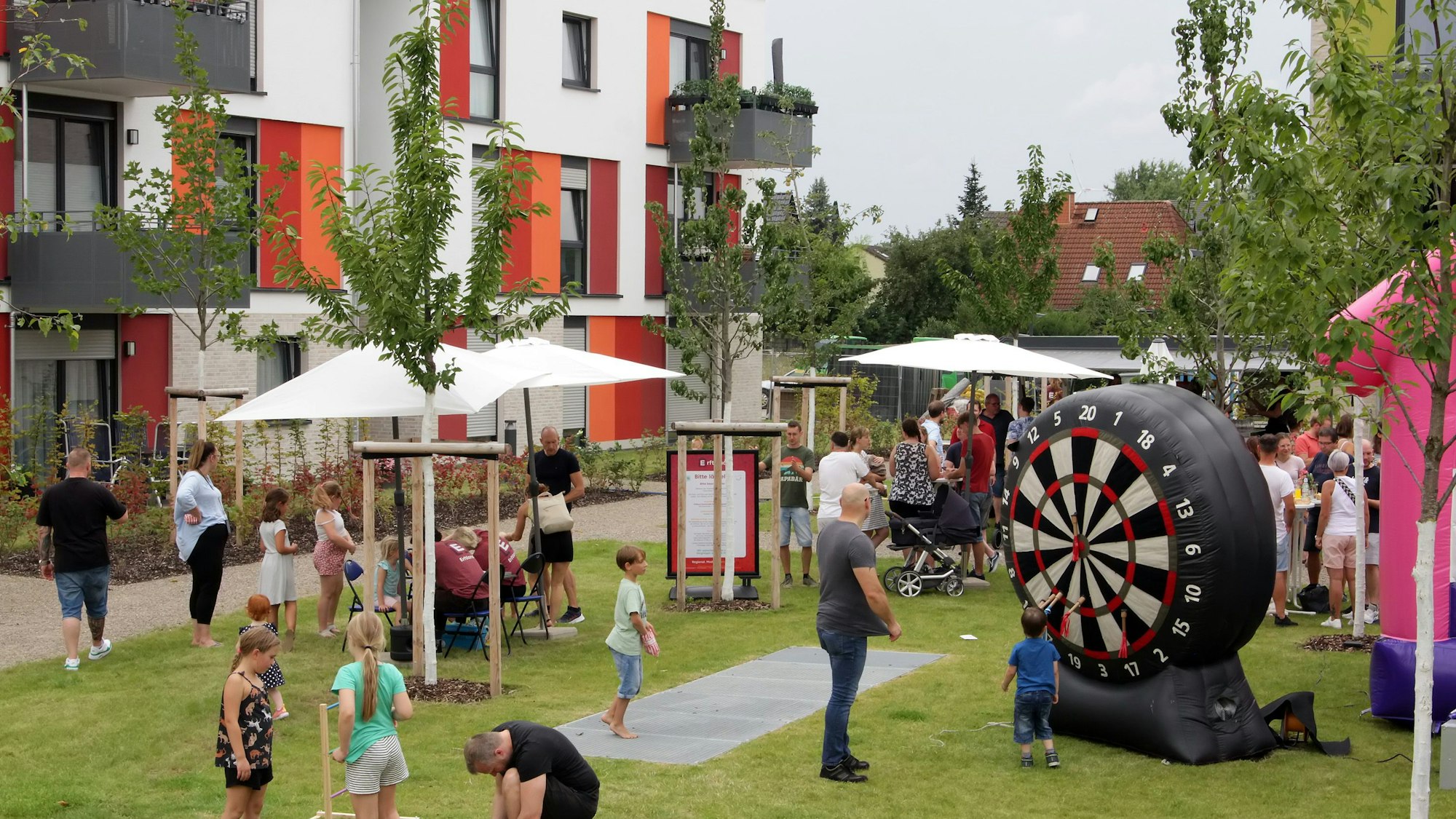 Auf einer WIese mit jungen Bäumen feiern Kinder und Erwachsene mit einer Hüpfburg und einer aufblasbaren Wurfscheibe, im Hintergrund steht ein neugebautes Mehrfamilienhaus.