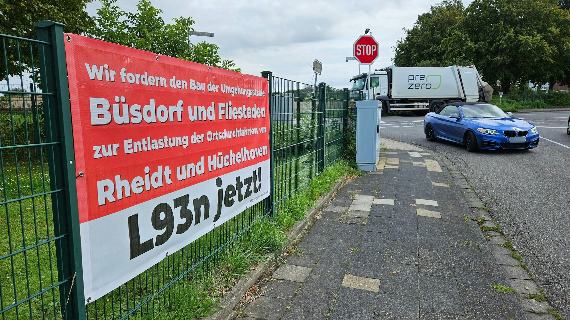 Ein rot-weißes Plakat, das an einem Zaun neben einer Straße befestigt ist, fordert den Bau einer Umgehungsstraße.