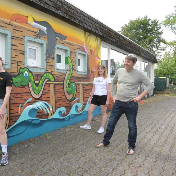 Vor einem Gebäude stehen zwei junge Mädchen und ein Mann. Die Fassade des Hauses wurde mit bunter Farbe bemalt. Das Motiv ist die Arche Noah.