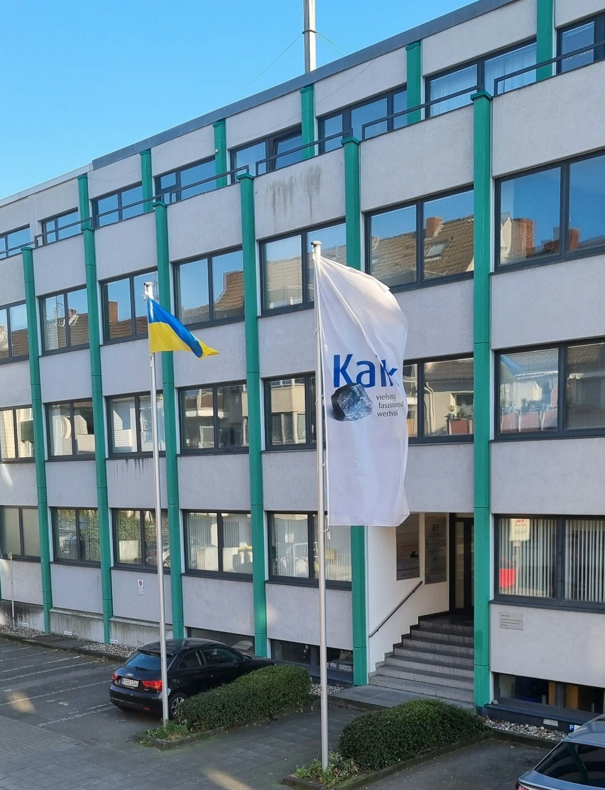 Ein mehrstöckiges Bürogebäude mit zwei Fahnenmasten und parkenden Autos ist zu sehen.