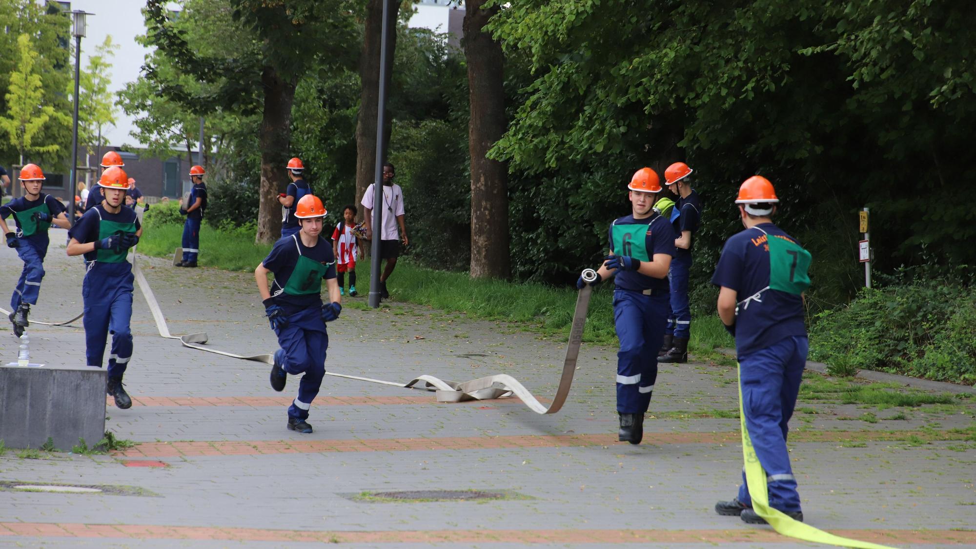 Jugendliche in Feuerwehrausrüstungen laufen über eine Straße und koppeln Schläuche aneinander.