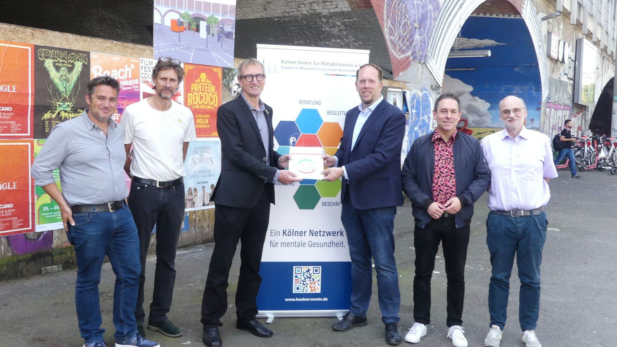 Vor einem Bahnbogen in Köln-Ehrenfeld stehen sechs Männer, die beiden in der Mitte halten gemeinsam einen Förderbescheid.
