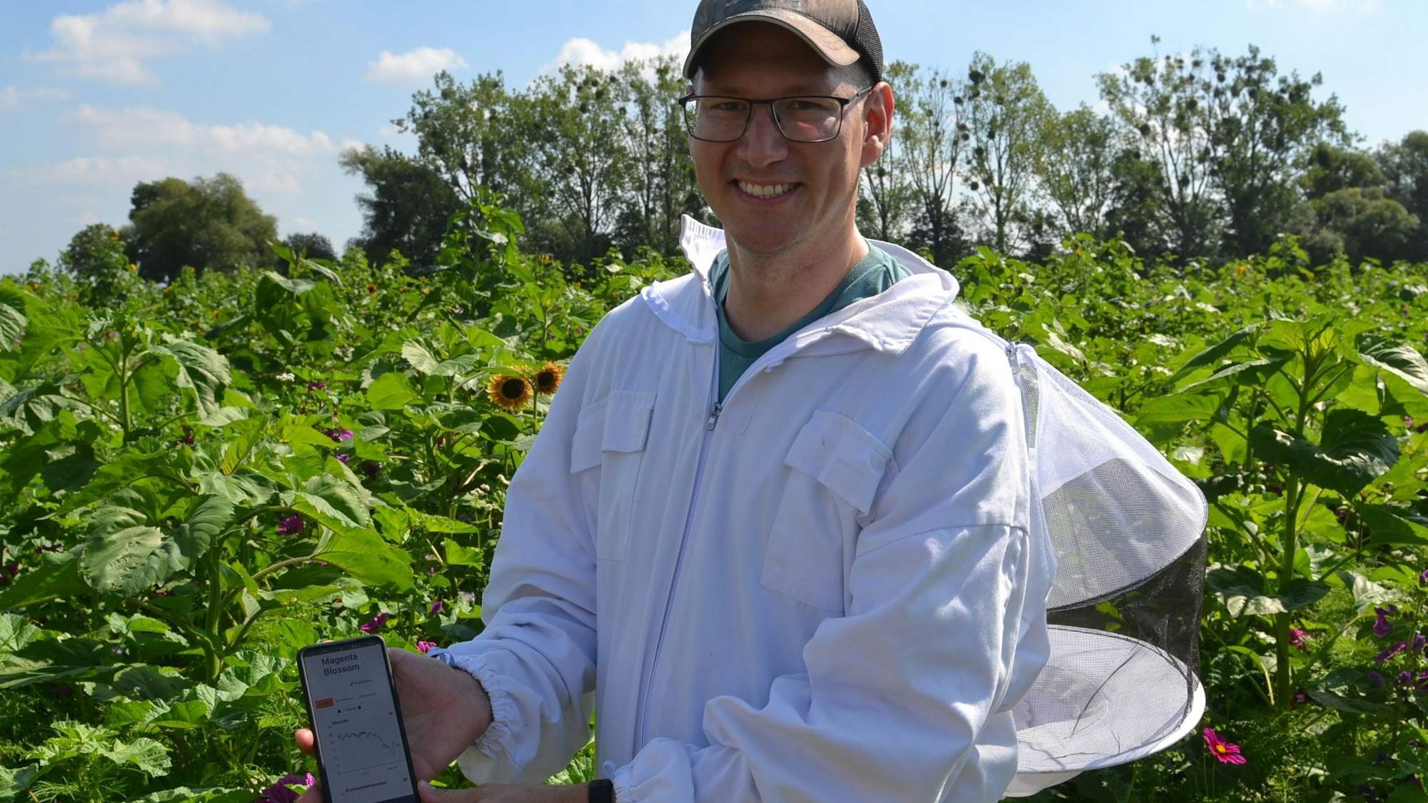 Der Imker Patrick Köhler trägt einen weißen Schutzanzug und hält ein Smartphone in der Hand. Über eine App den Gesundheitszustand der Bienen ablesen.