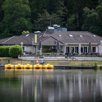Zwei Personen sitzen auf einer Bank am Tretbootverleih am Decksteiner Weiher, während im Hintergrund das Restaurant „Haus am See“ zu sehen ist.