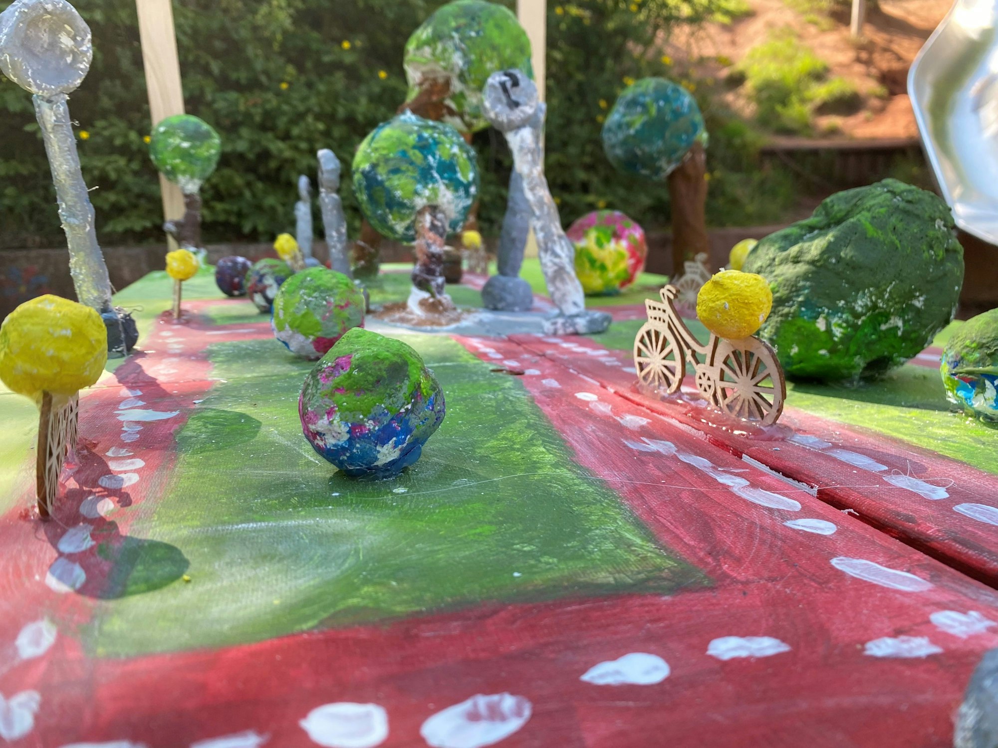 Auf dem Bild zu sehen ist ein buntes Pappmachémodell. Rote Wege sollen Fahrradstraßen darstellen. Darauf festgeklebt sind Holzschnitt-Fahrräder.