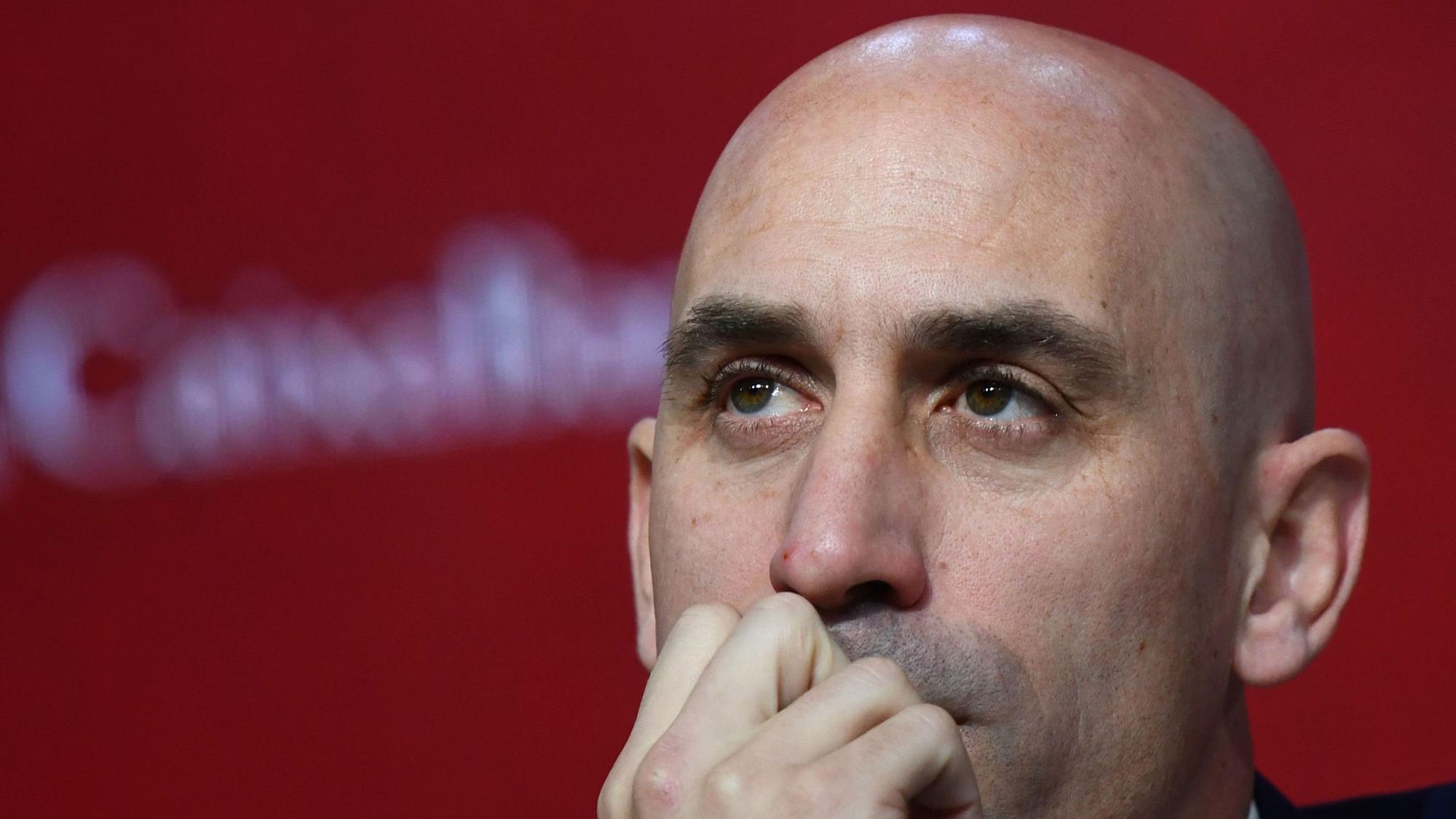Nahaufnahme von Luis Rubiales Gesicht vor einem unscharfen roten Hintergrund - Rubiales hält eine Hand am Mund und schaut in die Ferne.