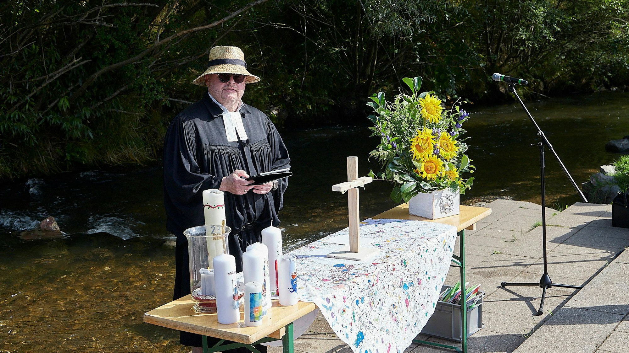 Pfarrer Oliver Joswig trägt Strohhut und Sonnenbrille. Er steht an einem Altar, der auf einem Holztisch vor der Olef in Hellenthal aufgebaut ist.