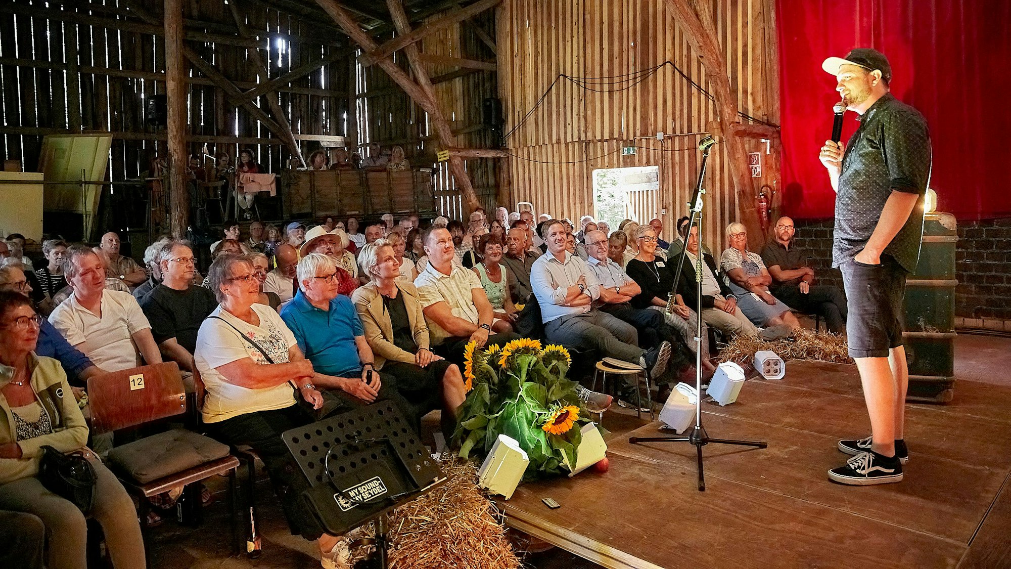 In Nettersheim wird eine alte Scheune im Sommer zur Kinoscheune. Auch eine Veranstaltung des Mundartfestivals fand dort statt - hier ist Poetry-Slammer Julius Esser auf der Bühne zu sehen, links und im Hintergrund das Publikum und die Szenerie der Bretterwand der urigen Scheune.