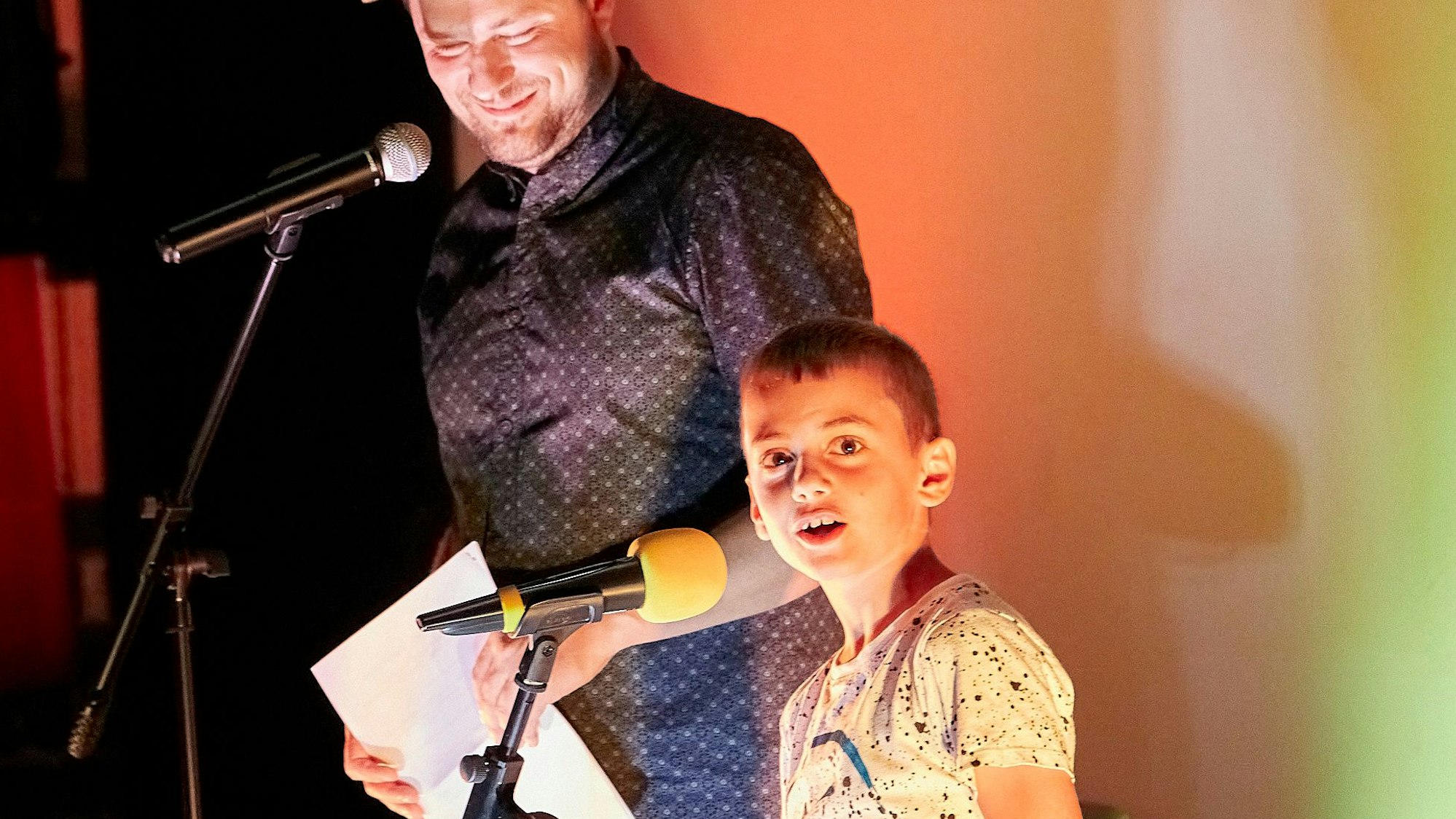 Der neunjährige Markus Hopfinger steht vor einem Mikrofon auf einer Bühne. Schräg hinter ihm steht der Poetry-Slammer Julius Esser.