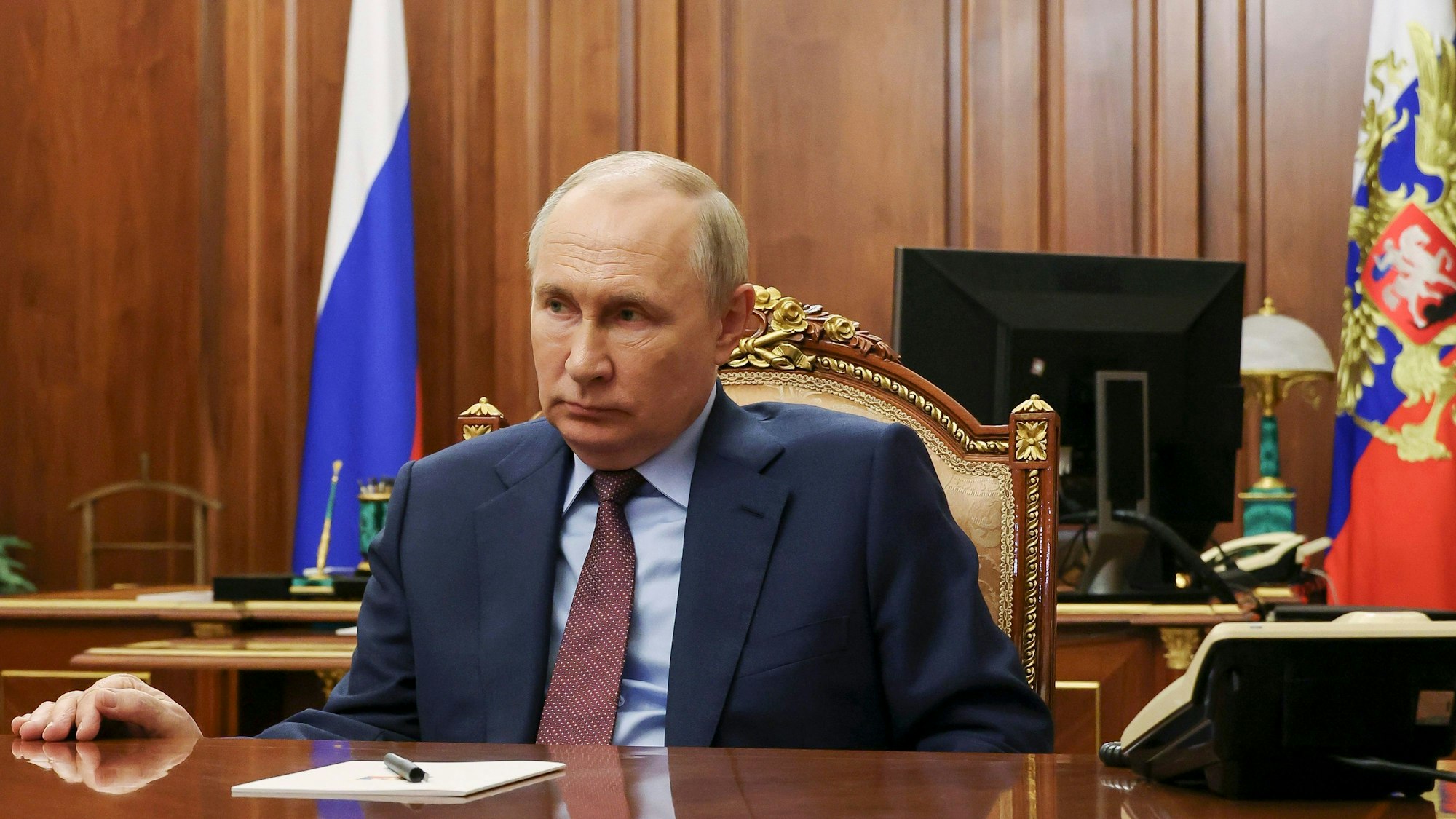 Der russische Präsident Wladimir Putin sitzt an einem Tisch in seinem Büro im Kreml. Im Hintergrund sind zwei russische Fahnen zu sehen.