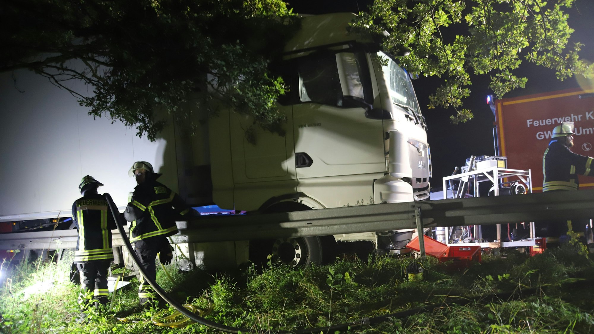 Nach dem Zusammenstoß eines Autos mit einem Pkw auf der A 3 kurz vor dem Ein weißer Lkw steht auf der Autobahn an einer Leitplanke, das Cockpit drückt die Leitplanke zur Seite. Feuerwehrleute stehen mit einem Schlauch daneben.