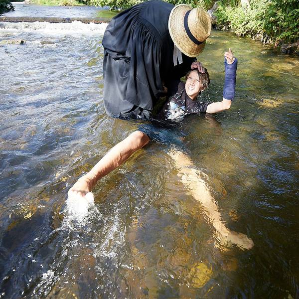Ein Mann mit Priestertalar und Strohhut hält einen Jungen bei der Taufe in der Olef im Wasser.