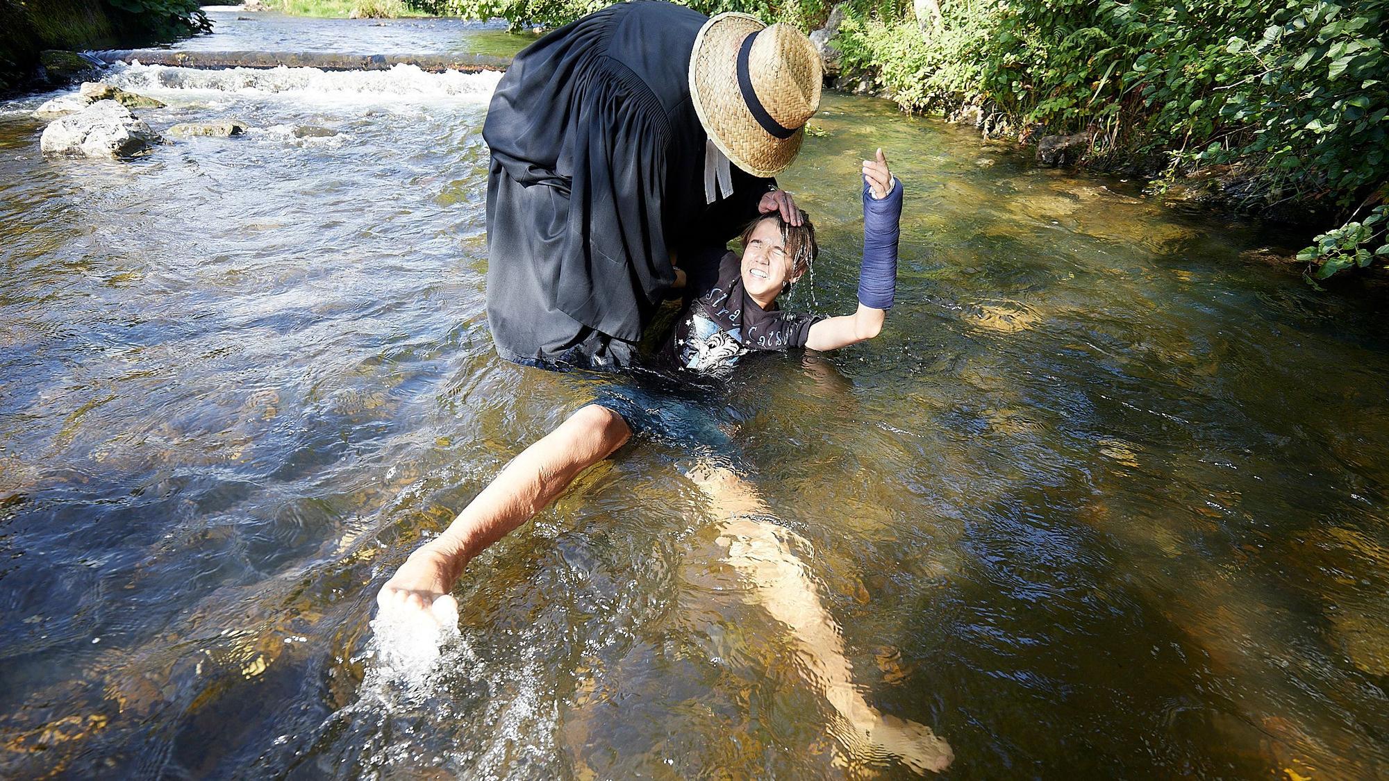 Ein Mann mit Priestertalar und Strohhut hält einen Jungen bei der Taufe in der Olef im Wasser.