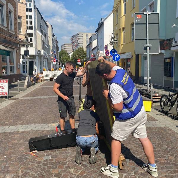 Das Bild zeigt Mitarbeiter der Stadt, die mit vereinten Kräften auf der Kölnstraße die Bodenplatte für eine Fahrradreparaturstation halten, um Schrauben anzubringen.