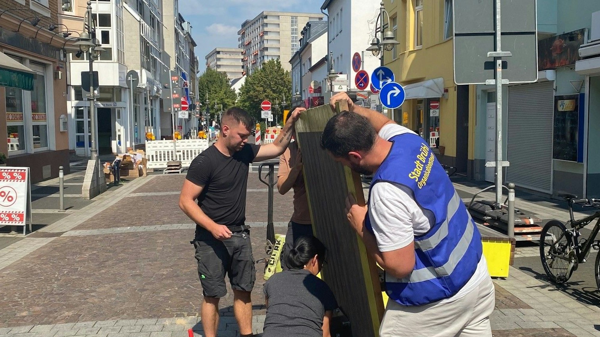 Das Bild zeigt Mitarbeiter der Stadt, die mit vereinten Kräften auf der Kölnstraße die Bodenplatte für eine Fahrradreparaturstation halten, um Schrauben anzubringen.