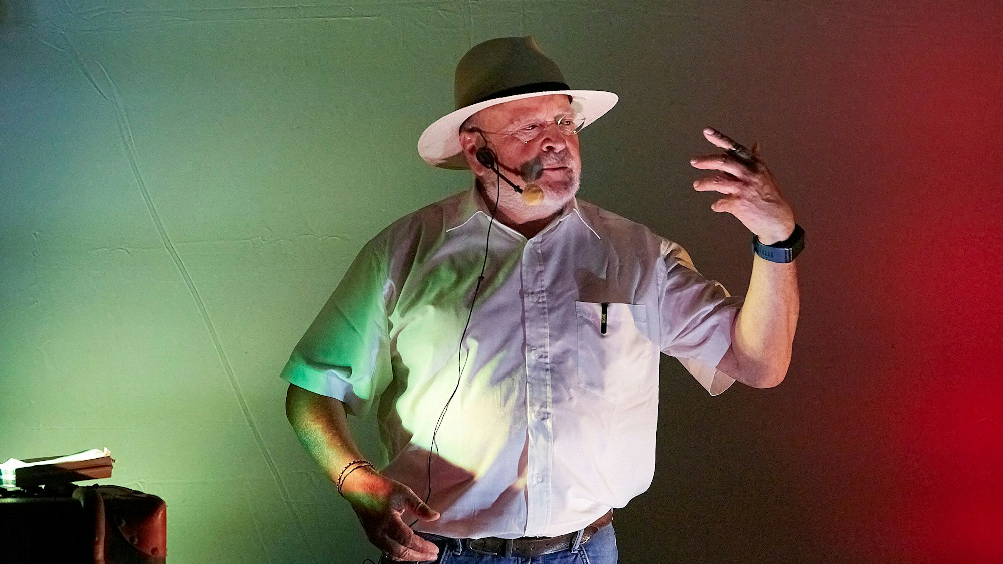 Der Journalist und Diakon Manfred Lang wird von rotem und grünem Bühnenlicht angestrahlt. Er hat ein Mikrofon vor dem Mund und berichtet beim Mundartfestival von den Feinheiten des Eifeler Platt.