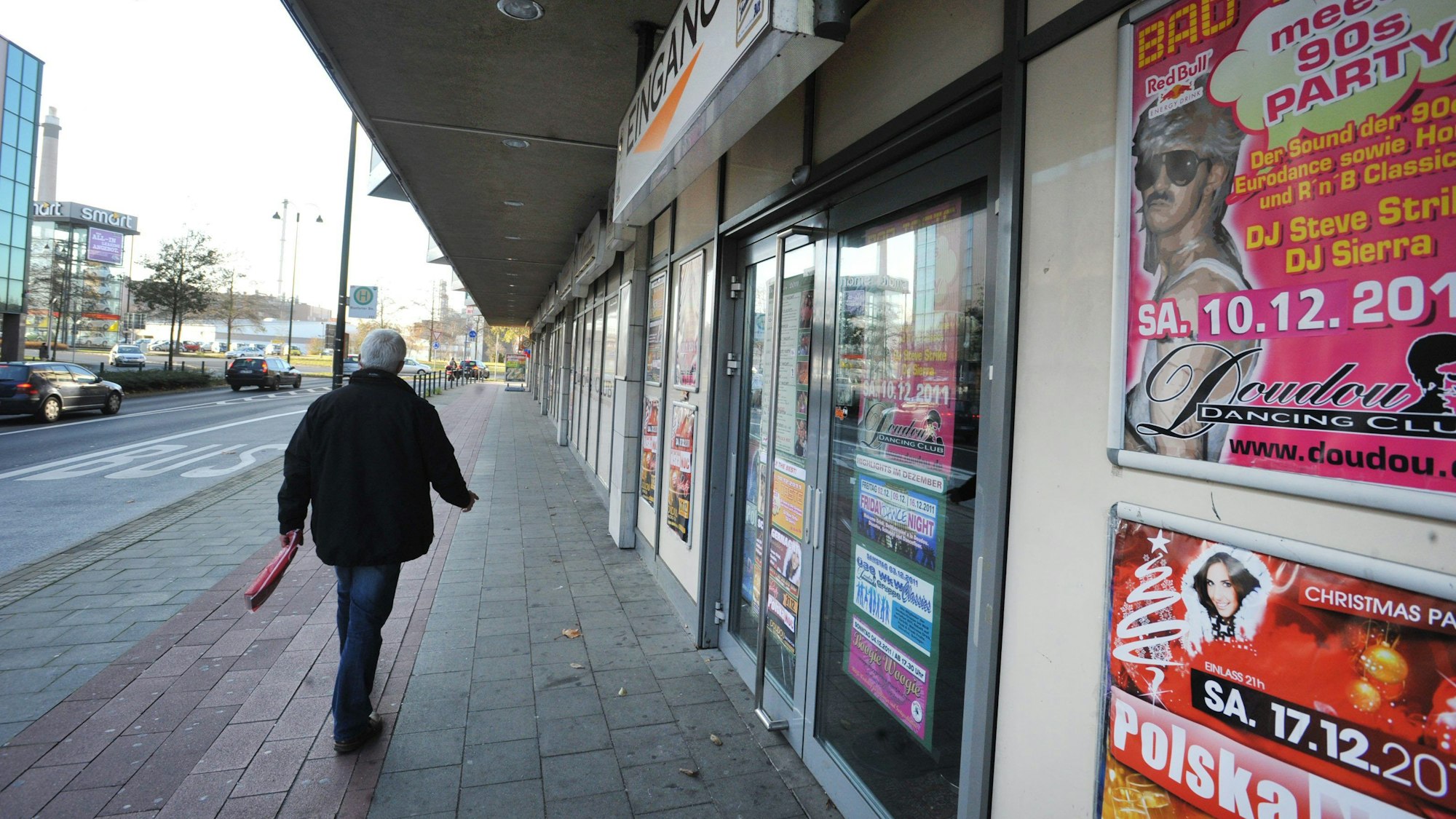 Ehemaliger Standort des Doudou Dancing Club in Leverkusen. Rechts sind Plakate an einer Wand aufgehängt, die auf Veranstaltungen hinweisen, links im Bild geht ein Mann über einen Fahrradweg.