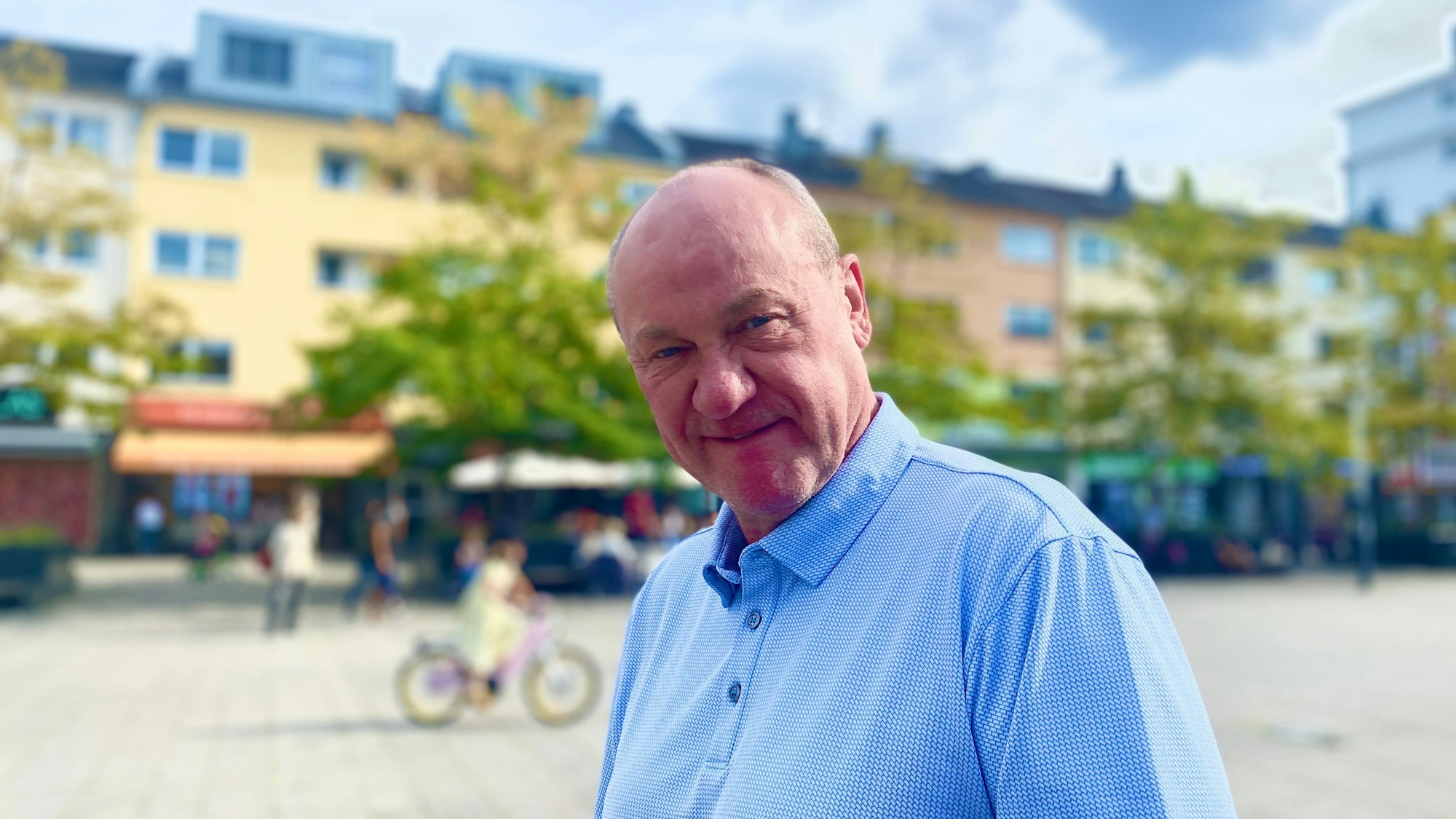 Ein mittelalter Mann mit wenigen Haaren und blauem Hemd steht auf einem Marktplatz.