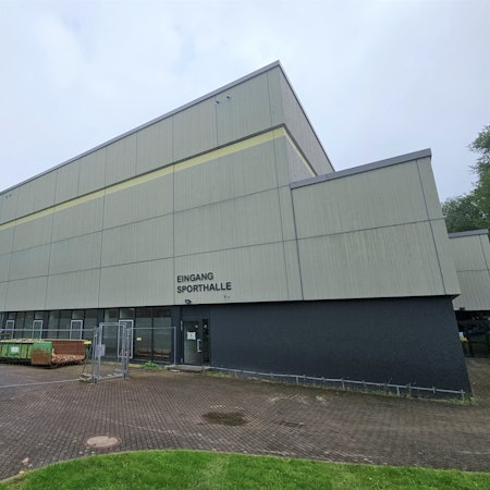 Das Bild zeigt die Sporthalle des Berufskollegs Eifel in Kall. Vor dem Gelände stehen Bauzäune und Container.