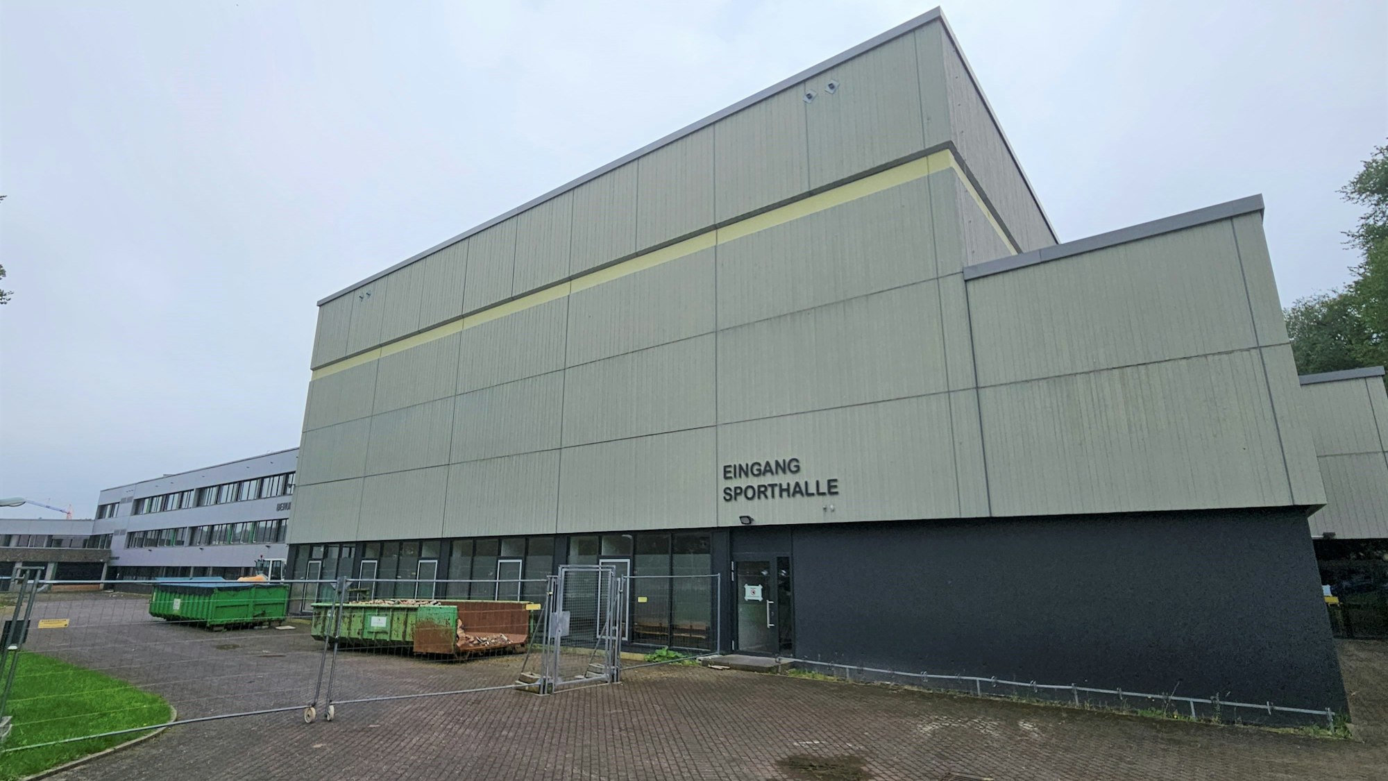 Das Bild zeigt die Sporthalle des Berufskollegs Eifel in Kall. Vor dem Gelände stehen Bauzäune und Container.