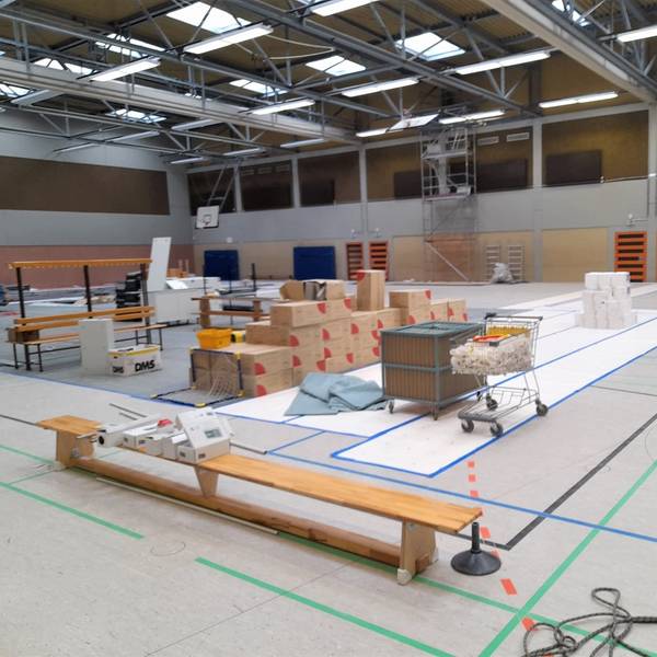 Das Bild zeigt eine Turnhalle. Auf einer Sportbank liegen Pakete. Große Kartons sind in der Mitte der Halle gelagert. Ein Einkaufswagen ist mit Plastik befüllt. An der Wand der Tundhalle steht ein Gerüst.