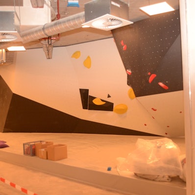 Auf schwarzen und weißen Wandelementen sind bunte Griffe festgeschraubt. An der Decke verlaufen die Rohre der Lüftungsanlage. Baustellenband sperrt den Zugang ab.