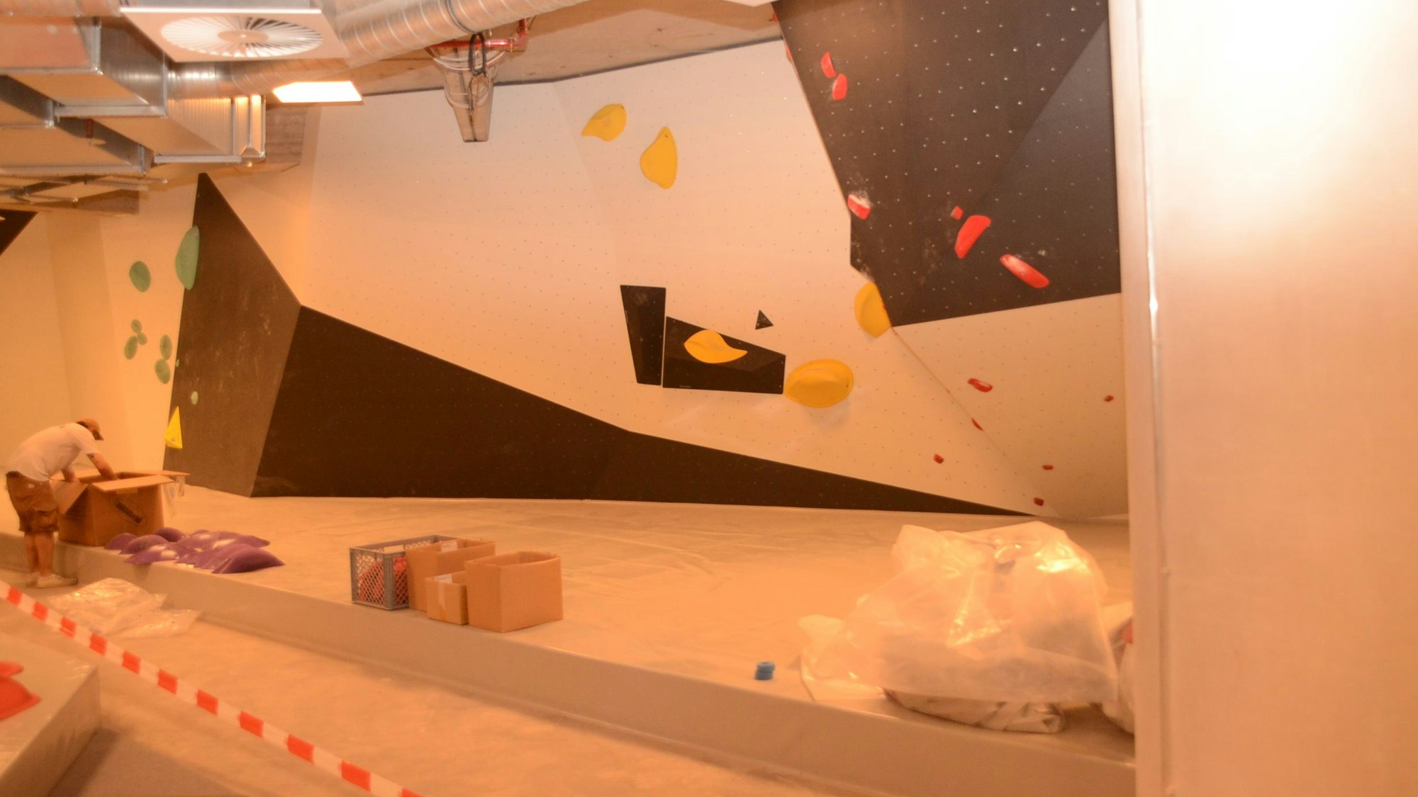 Auf schwarzen und weißen Wandelementen sind bunte Griffe festgeschraubt. An der Decke verlaufen die Rohre der Lüftungsanlage. Baustellenband sperrt den Zugang ab.