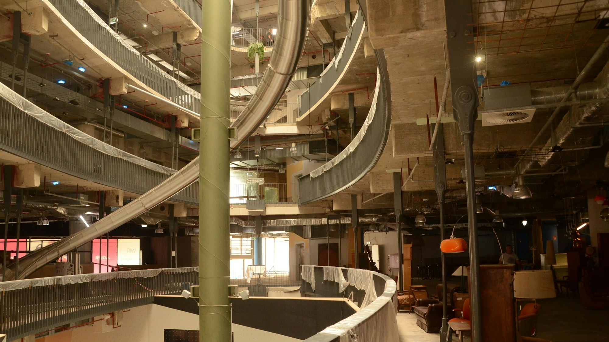 Eine ovale Öffnung erstreckt sich über mehrere Etagen, eine Rutsche aus Edelstahl führt im Bogen herunter. Rechts stehen alte Möbel für die Inneneinrichtung.