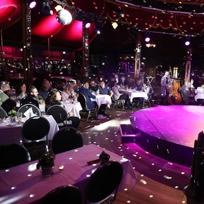 Ein Künstler steht auf der runden Bühne eines Spiegelzelts in Altenberg und bereitet seinen Auftritt vor. Auf Stühlen an Tischen sitzen Gäste.