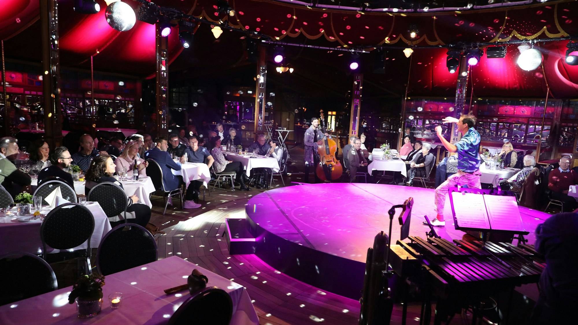 Ein Künstler steht auf der runden Bühne eines Spiegelzelts in Altenberg und bereitet seinen Auftritt vor. Auf Stühlen an Tischen sitzen Gäste.