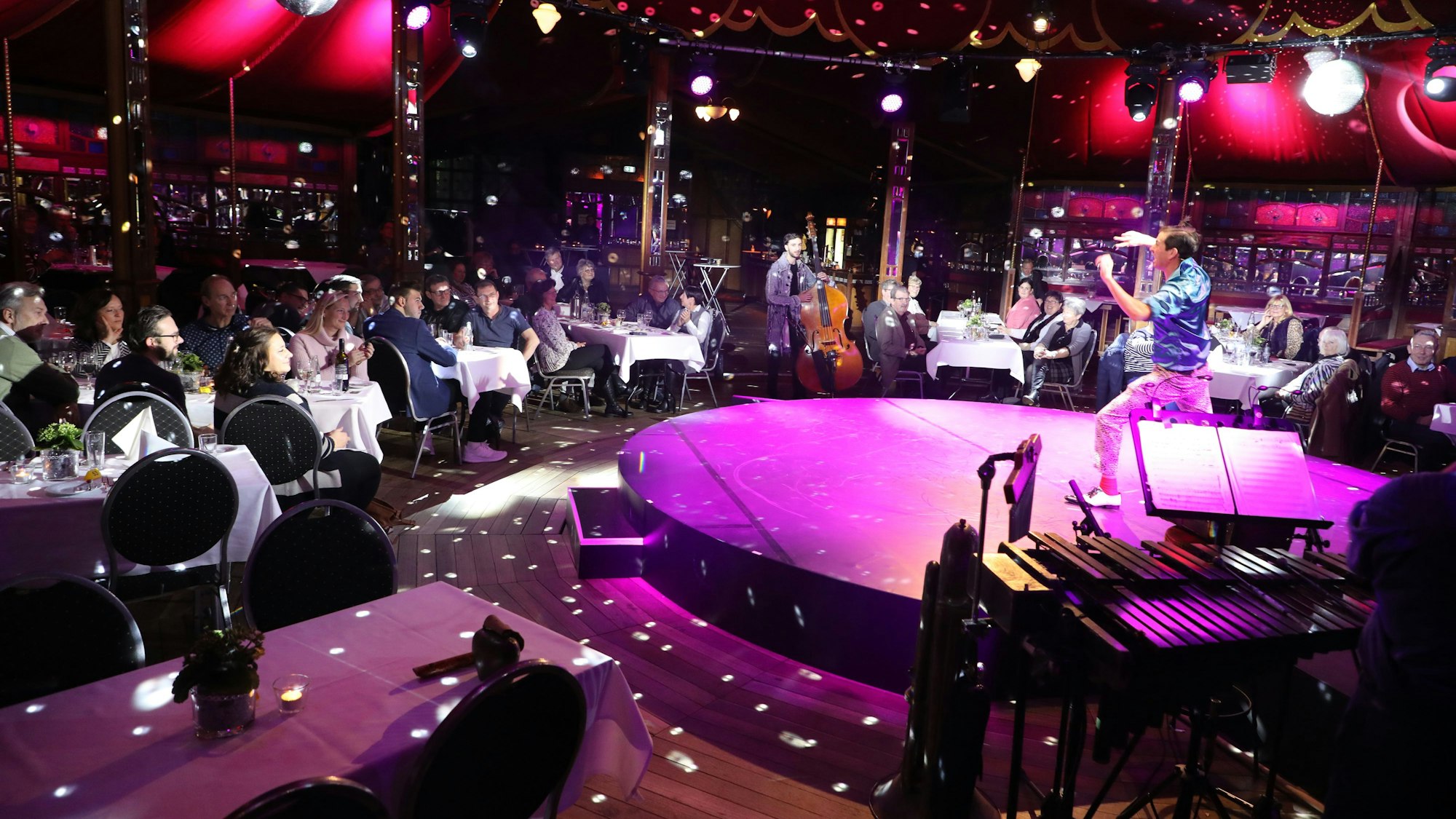 Ein Künstler steht auf der Bühne des Altenberger Spiegelzelts. Menschen sitzen an Tischen und schauen zu.