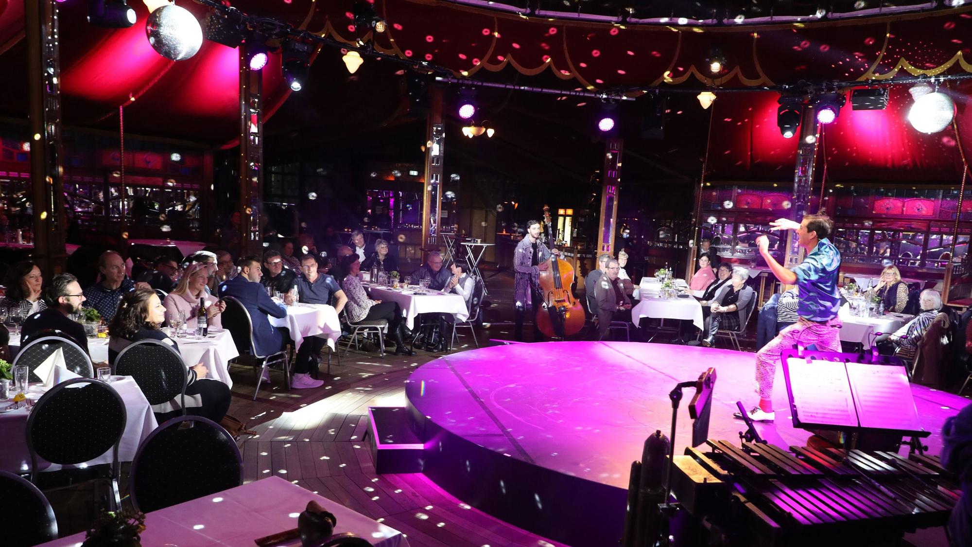 Ein Künstler steht auf der Bühne im Altenberger Spiegelzelt. Menschen sitzen an Tischen bei der Dinnershow mit dem Kölner Cirque Bouffon.