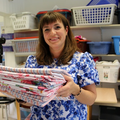 Das Bild zeigt die Ukrainerin Tatjana Jezmakova mit einem Stapel frisch gewaschener Bettlaken in ihrer Reinigung. Hinter ihr stehen viele Körbe für die Wäsche.