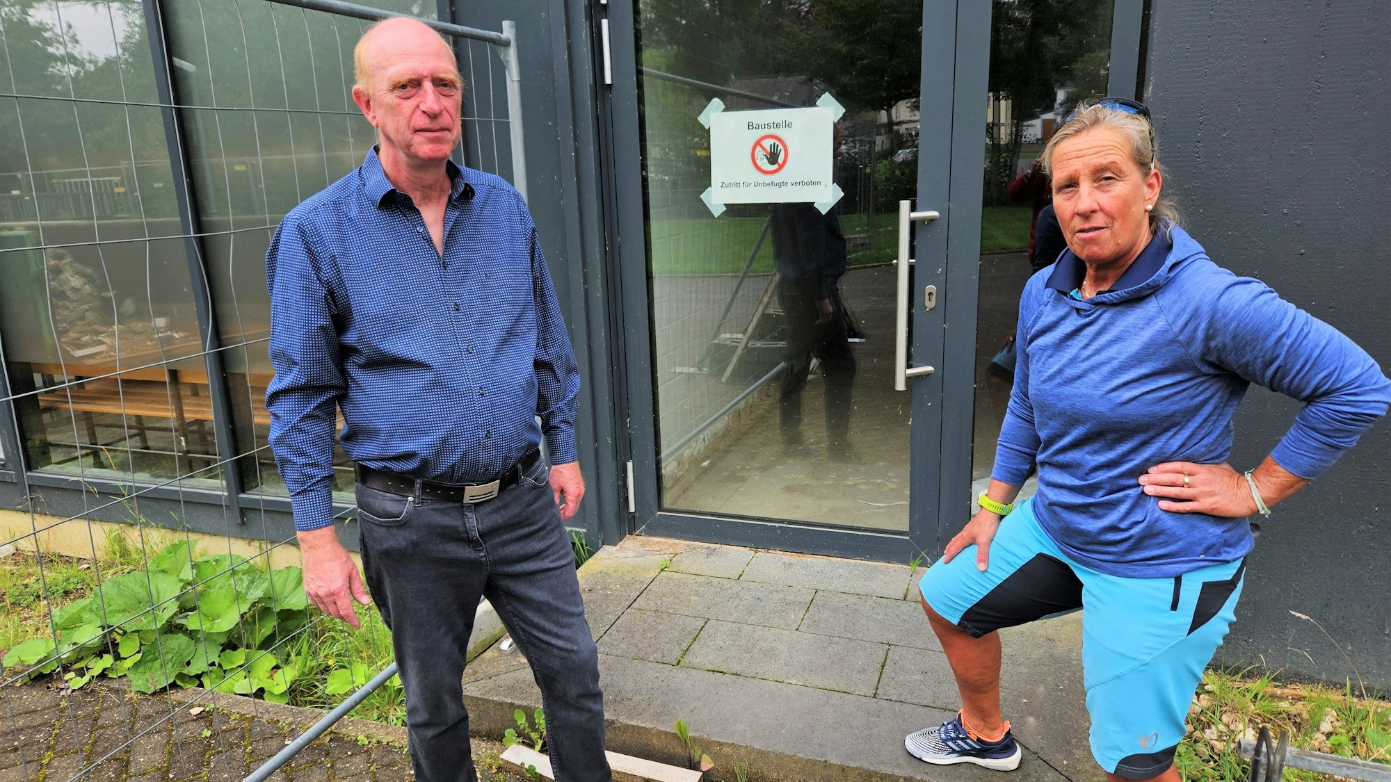 Vor dem Eingang der Halle stehen Richard Fremery und Iris Schröder vom TV Kall. An der Eingangstür hängt ein Warnschild mit der Aufschrift „Baustelle“. Neben der Tür: ein Bauzaun.