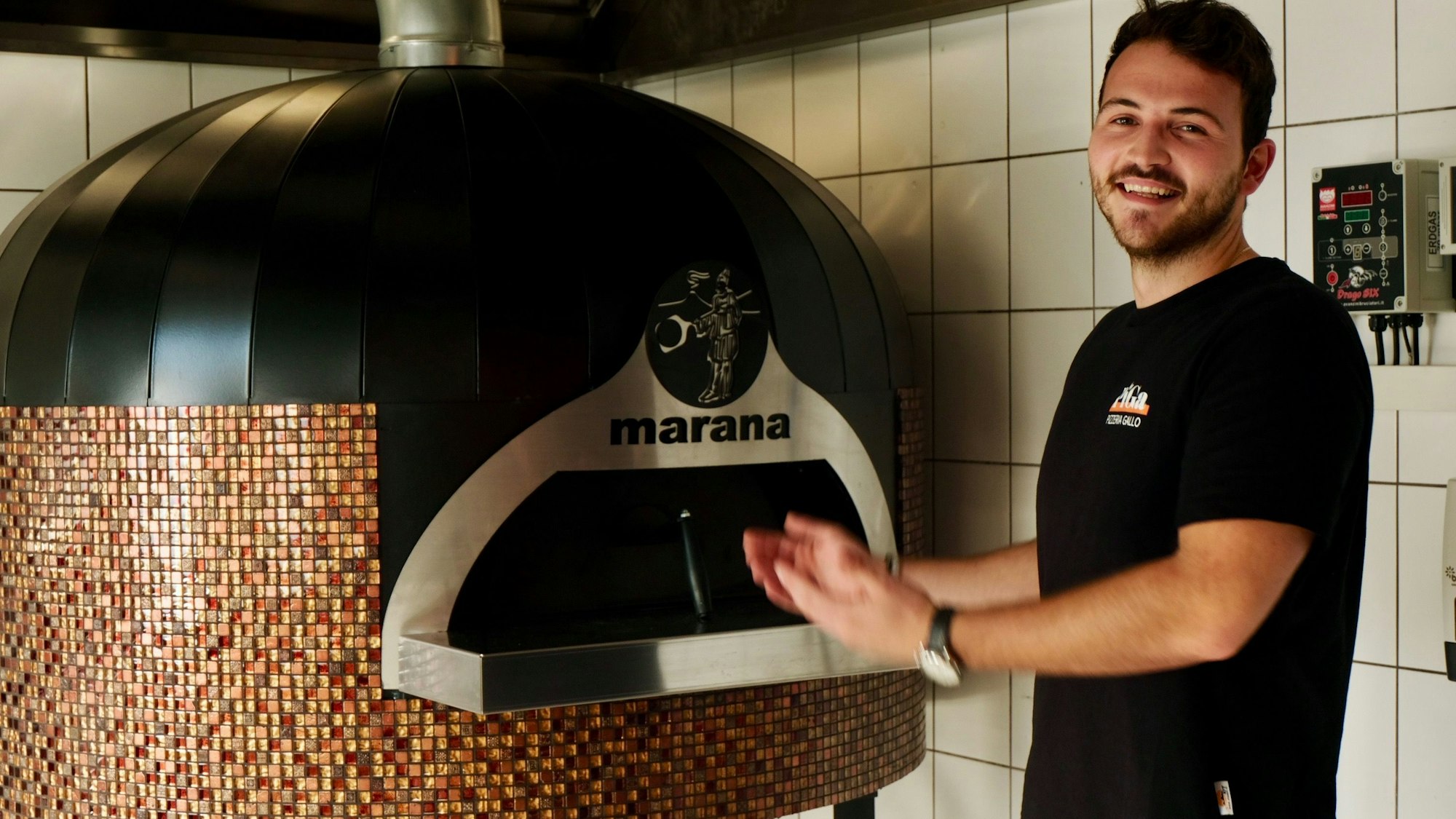 Ein junger Mann mit braunem Haar und Bart steht neben einem runden mosaik-besetzten Ofen.