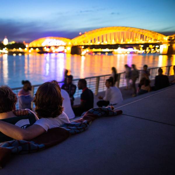 Menschen sitzen bei Sonnenuntergang auf dem Rheinboulevard am Rhein in Köln (Nordrhein-Westfalen) mit Blick auf den Dom.
