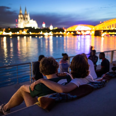 Menschen sitzen bei Sonnenuntergang auf dem Rheinboulevard am Rhein in Köln (Nordrhein-Westfalen) mit Blick auf den Dom.