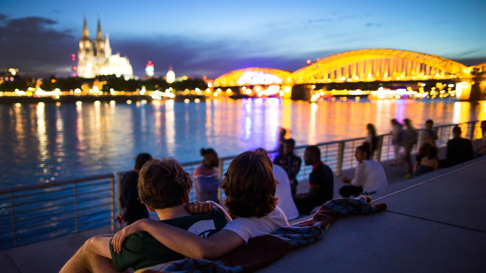 Menschen sitzen bei Sonnenuntergang auf dem Rheinboulevard am Rhein in Köln (Nordrhein-Westfalen) mit Blick auf den Dom.