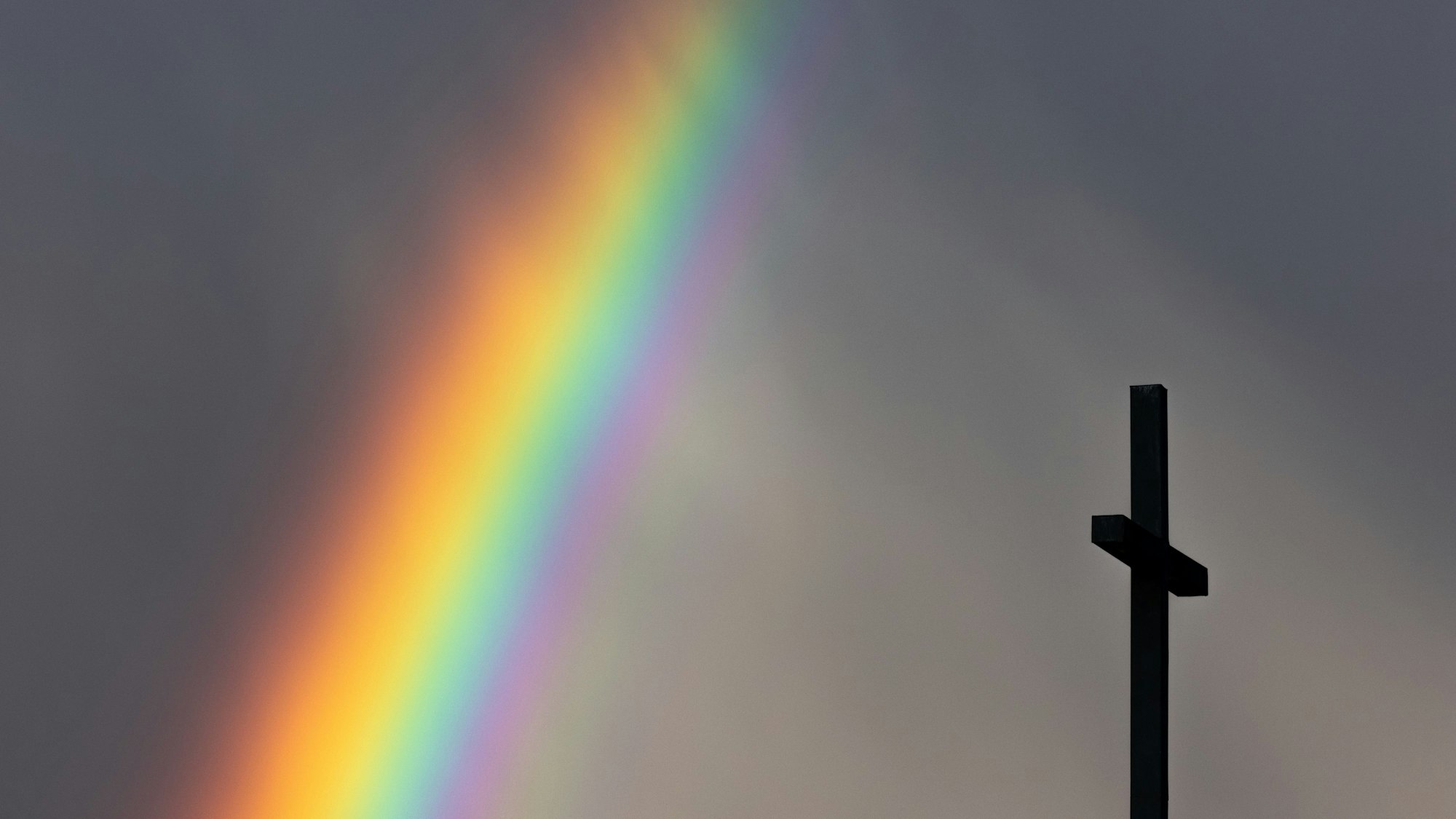 Ein kräftiger Regenbogen ist am dunklen Himmel neben dem Kreuz einer Kirche zu sehen. Die evangelische Kirche plant ein Popup-Hochzeitsfest an der Christus-Kirche. „Vielfalt feiern“ ist das Motto, so der evangelische Kirchenverband.