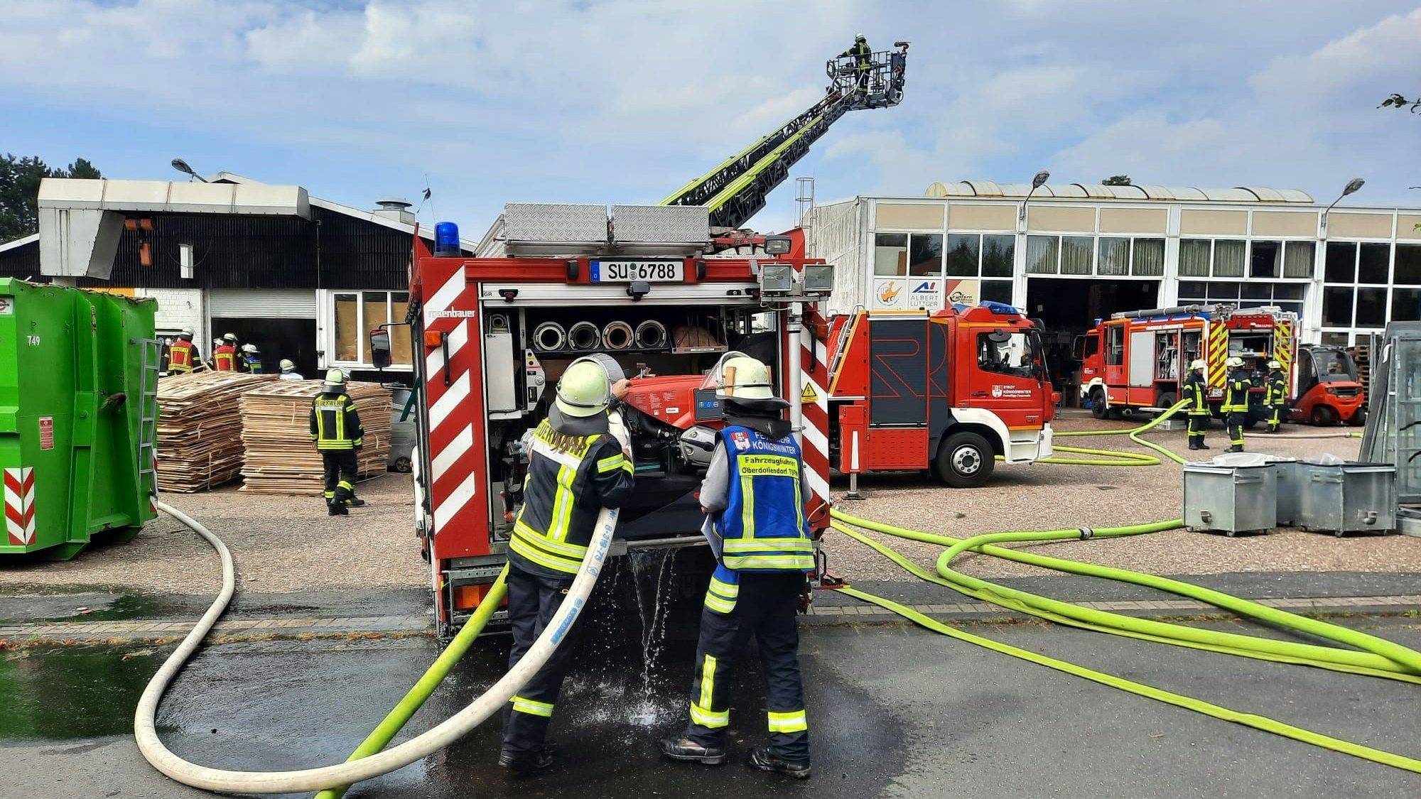 Einsatzfahrzeuge der Feuerwehr stehen vor einem Betriebsgebäude. Auf der Fahrbahn liegen gelbe Schlauchleitungen. Die Drehleiter ist ausgefahren.