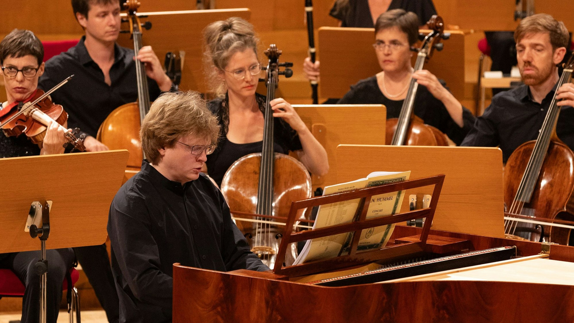 Der Pianist sitzt an einem Hammerklavier, hinter ihm eine Reihe Cellisten.
