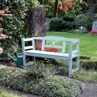 Eine weiße Holzbank steht neben einem Grab auf dem Kölner Melaten-Friedhof.