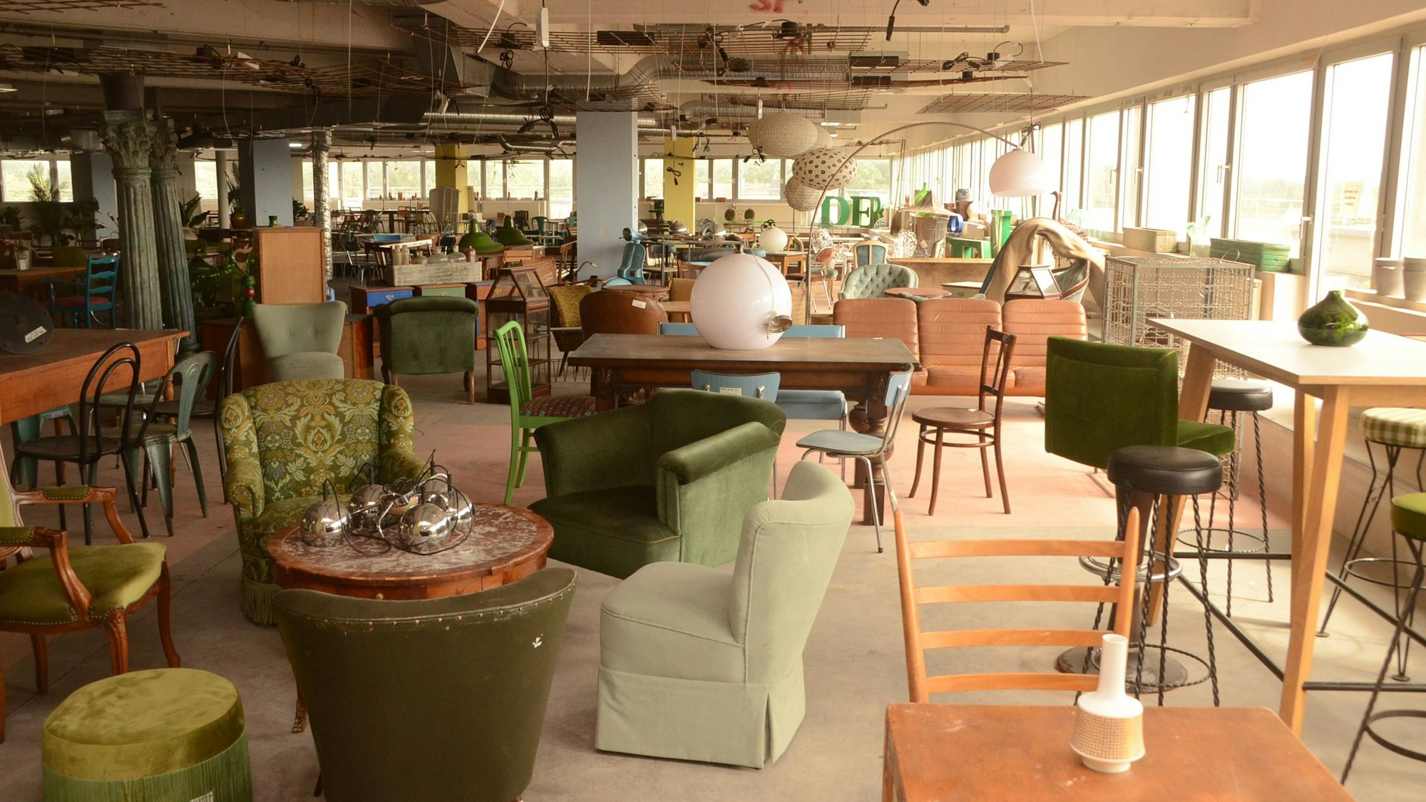 Moderne und altmodische Möbel in einem Raum mit umlaufenden Glasfenstern. Die Decke ist nicht verkleidet, man sieht Kabel und andere Technik.