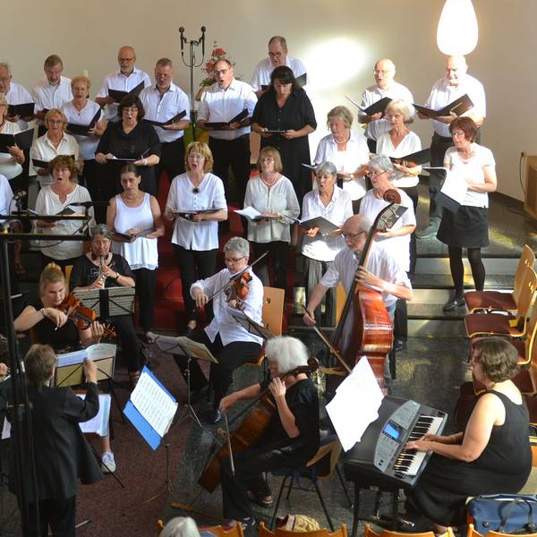 Das Bild zeigt einen Kirchenchor. Im Vordergrund sitzen Musiker mit Cello, Geigen, E-Piano und Flöten. Im Hintergrund stehen die Sänger und Sängerinnen.