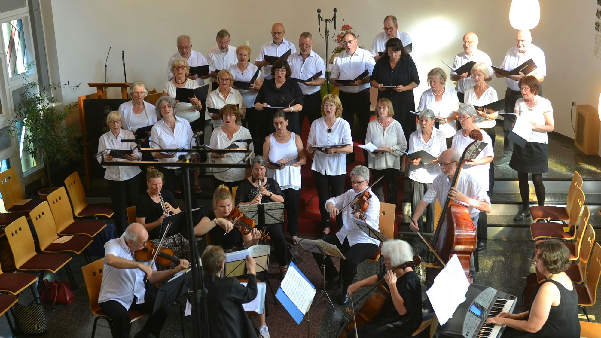 Das Bild zeigt einen Kirchenchor. Im Vordergrund sitzen Musiker mit Cello, Geigen, E-Piano und Flöten. Im Hintergrund stehen die Sänger und Sängerinnen.