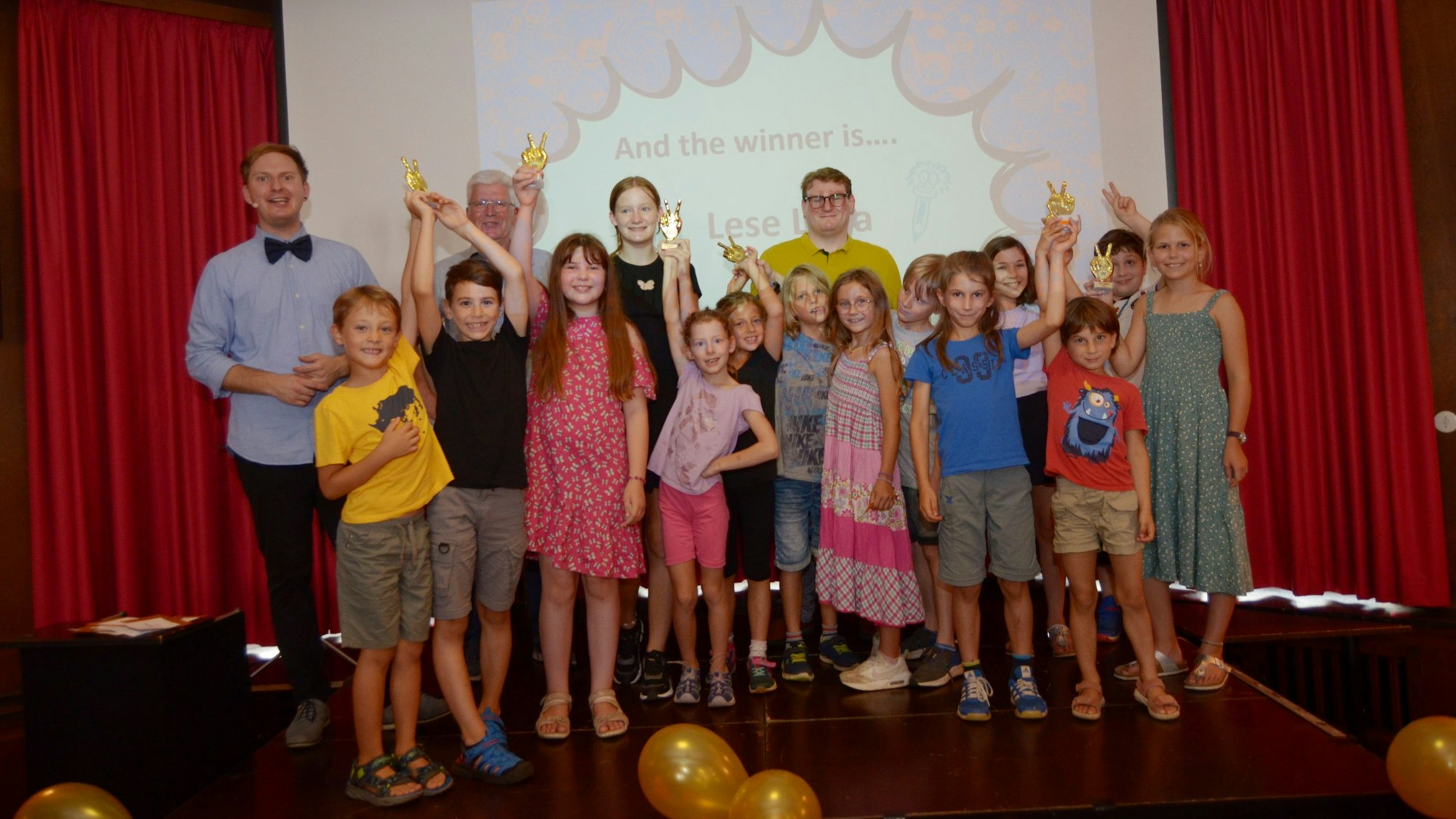 Auf der Bühne im Euskirchener Casino stehen die Kinder und Jugendlichen, die beim Abschlussfest des Sommerleseclubs der Stadtbibliothek als Auszeichnung „Lese-Oskars“ erhalten haben. Einige halten ihre Trophäen in die Höhe.