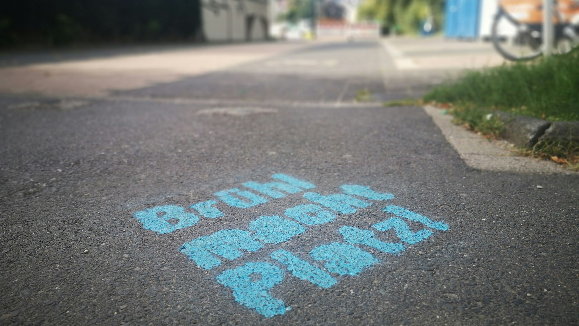 Die Worte „Brühl macht Platz!“ in blauer Farbe auf dem Boden. Im Hintergrund der Belvedere-Parkplatz völlig ohne Autos.