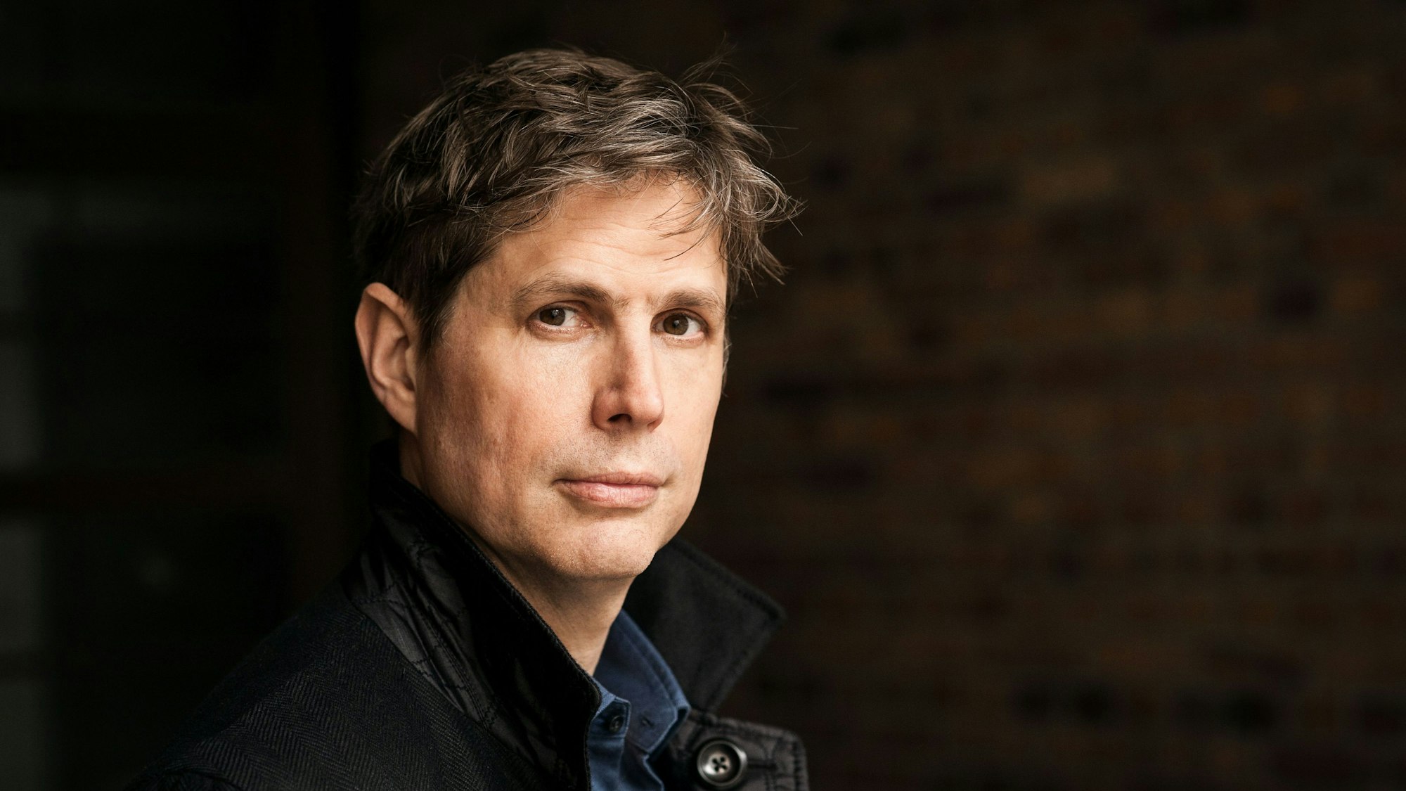 Daniel Kehlmann steht vor einer dunklen Wand. Er trägt eine dunkle Jacke und blickt in die Kamera.