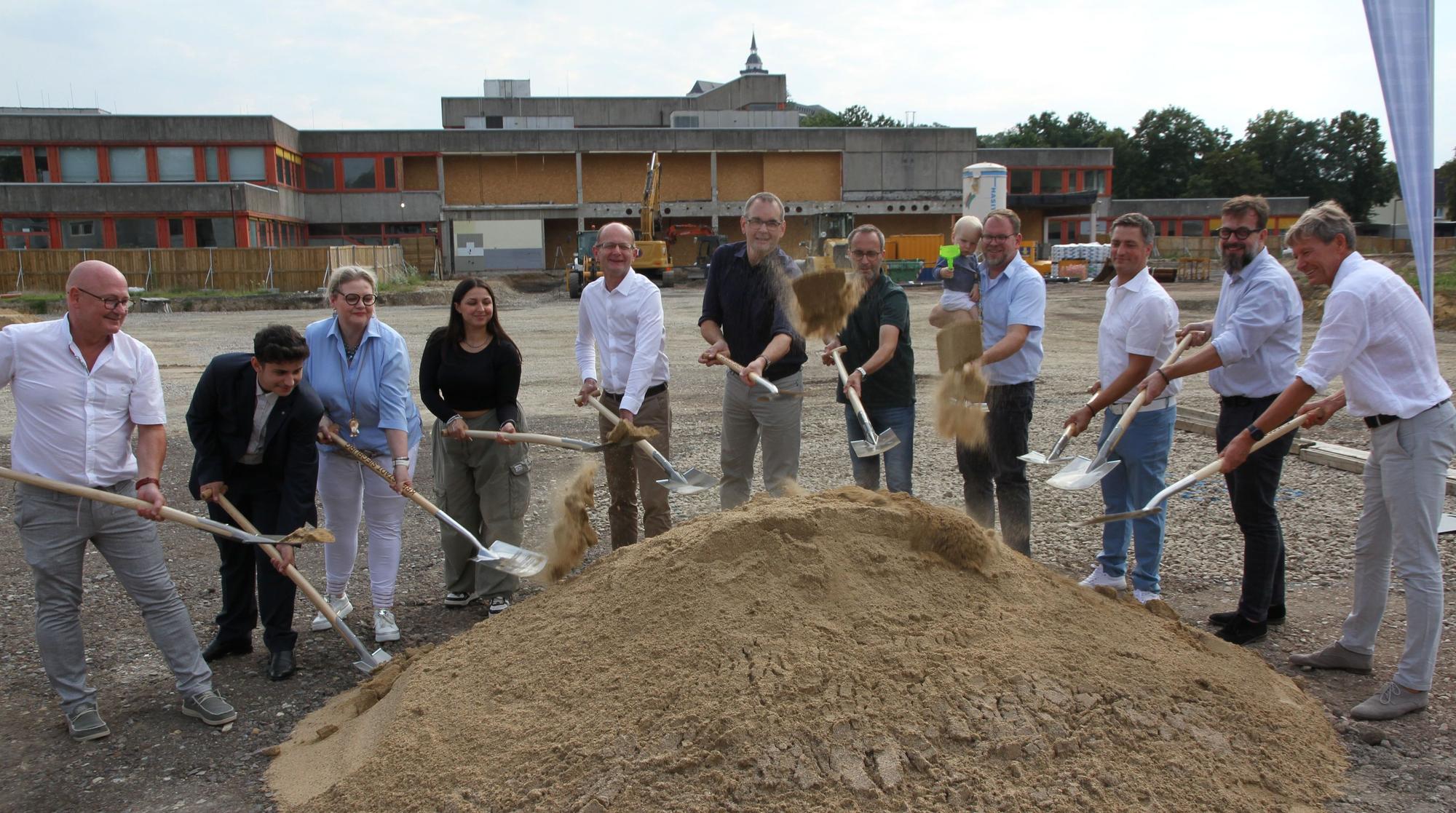 Zwölf Menschen schaufeln für einen Spatenstich Sand aus einem Sandhaufen.