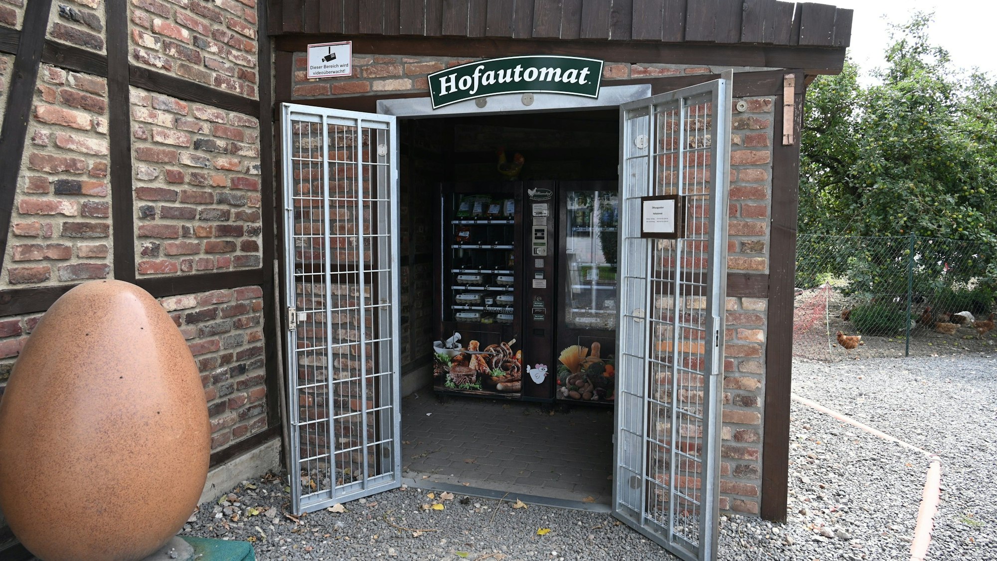 Ein Anbau mit geöffneten Türen. Durch den Eingang kann man den Hofautomaten entdecken. Darüber ein Schild mit der Aufschrift „Hofautomat“. Im Vordergrund ist ein riesiges Ei zu sehen.